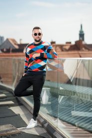 Ein Mann mit Sonnenbrille, einem bunten Argyle-Pullover und schwarzen Hosen lehnt an einem Glasgeländer auf einem Dach. Im Hintergrund sind Stadtdächer unter einem klaren Himmel zu sehen.