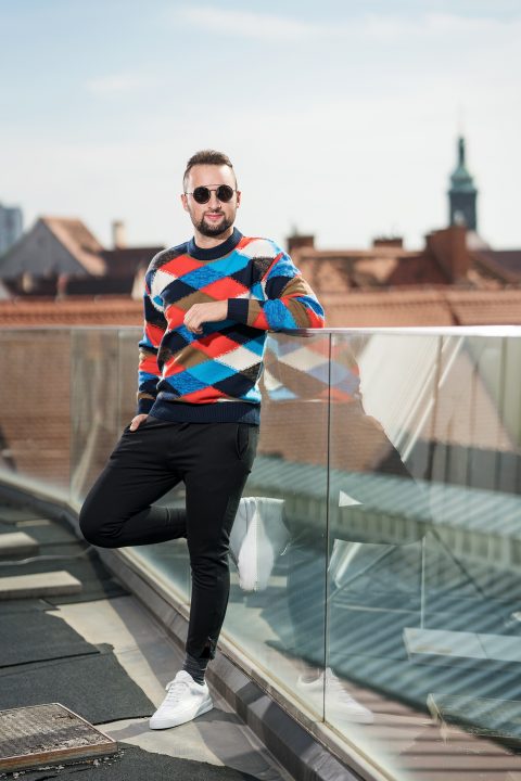 Ein Mann mit Sonnenbrille, einem bunten Argyle-Pullover und schwarzen Hosen lehnt an einem Glasgeländer auf einem Dach. Im Hintergrund sind Stadtdächer unter einem klaren Himmel zu sehen.