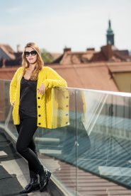 Eine Frau in einer leuchtend gelben Strickjacke, einem schwarzen Oberteil und schwarzen Hosen lehnt an einem Glasgeländer auf einem Dach. Sie trägt eine Sonnenbrille. Im Hintergrund sind eine verschwommene Stadtlandschaft und Dächer zu sehen.