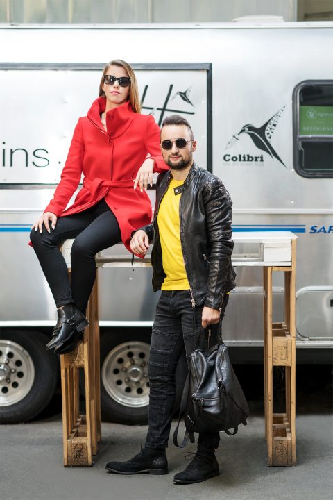 Zwei stylische Personen posieren selbstbewusst im Freien. Einer sitzt mit rotem Mantel und Sonnenbrille auf einem Tisch, der andere steht daneben, trägt eine schwarze Lederjacke und ein gelbes Hemd und hält einen Rucksack. Sie stehen vor einem Metallanhänger.