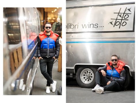 Eine Person mit einer auffälligen, farbenfrohen Jacke und Sonnenbrille lehnt links an einer reflektierenden Oberfläche. Rechts sitzt sie auf dem Boden neben einem metallischen Fahrzeug, im Hintergrund steht der Text „Colibri gewinnt ...“.