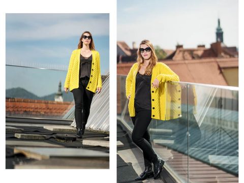 Eine Frau steht auf einem Dach und trägt eine leuchtend gelbe Strickjacke, ein schwarzes Oberteil, schwarze Hosen und eine Sonnenbrille. Der Hintergrund zeigt einen klaren Himmel und Dächer mit Gebäuden in der Ferne.
