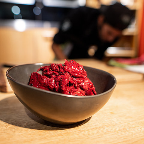 Eine schwarze Schüssel mit leuchtend rotem Gelato steht auf einem Holztisch. Im unscharfen Hintergrund beugt sich eine Person mit schwarzer Mütze und schwarzem Outfit vor und scheint in einer Küche Essen zuzubereiten.