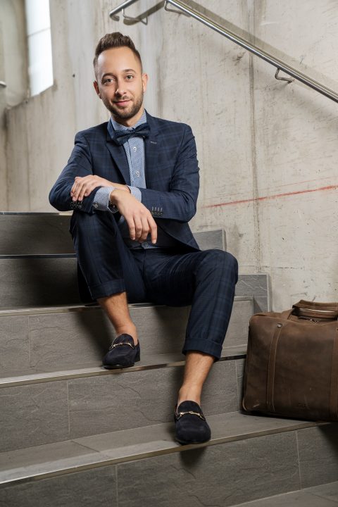 Ein Mann im karierten Anzug sitzt mit übereinandergeschlagenen Beinen auf einer Innentreppe. Er trägt eine Fliege und Slipper und hat einen Arm auf seinem Knie abgelegt. Neben ihm steht eine braune Ledertasche. Die Umgebung wirkt lässig und modern.