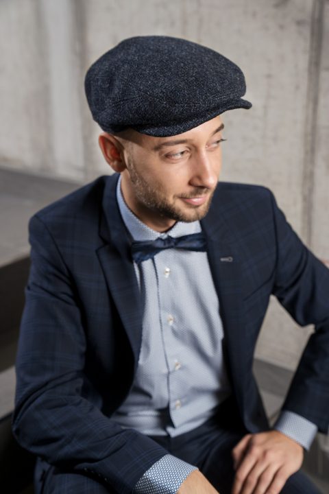 Ein Mann mit Bart trägt eine dunkle Schiebermütze, einen karierten Anzug, ein hellblaues Hemd und eine Fliege. Er sitzt und blickt mit neutralem Gesichtsausdruck vor einem Betonhintergrund zur Seite.