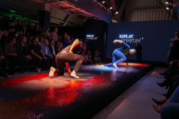 Zwei Darsteller tanzen energisch auf einem mit Glitzer bedeckten Laufsteg, umgeben von einem Publikum. Der Hintergrund ist mit dem „Replay Hyperflex+“-Branding versehen. Die Beleuchtung erzeugt bunte Reflexe auf dem Boden.