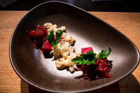 Ein Gourmetgericht wird elegant auf einem dunklen, asymmetrischen Teller serviert. Es besteht aus rotem gewürfeltem Fleisch, weißen Garnituren und grünen Kräutern, begleitet von einem Klecks cremiger Soße, kunstvoll arrangiert auf einer Holzoberfläche.
