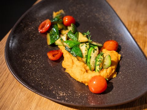 Auf einem dunklen, dreieckigen Teller liegt ein cremiges Orangenpüree, darauf sautierte Zucchini und Kirschtomaten, garniert mit frischen Kräutern. Das Gericht ist elegant auf einem Holztisch angerichtet.