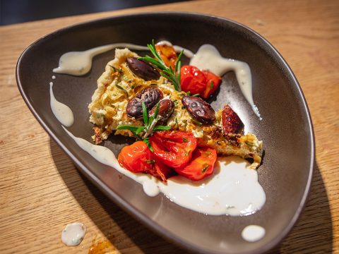 Ein Stück pikante Torte, garniert mit Kirschtomaten, Oliven und Rosmarinzweigen, wird auf einem dunklen, ovalen Teller präsentiert. Die Torte ist mit einer cremigen Sauce beträufelt und ruht auf einer Holzoberfläche.