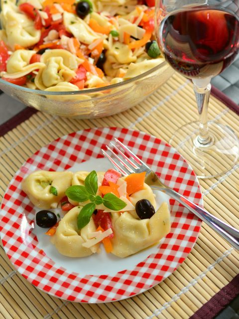 Ein Teller Tortellinisalat mit Oliven, Paprika und Basilikum auf einem rotkarierten Teller. Neben dem Teller steht ein Glas Rotwein, im Hintergrund eine größere Schüssel Salat. Auf dem Teller liegt eine Gabel.