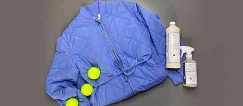 Eine blaue Steppjacke liegt flach auf einem grauen Untergrund, daneben drei gelbe Tennisbälle. Daneben stehen zwei Flaschen mit Textilpflegemitteln: eine mit Sprühkopf, die andere mit Schraubverschluss.