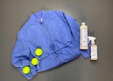 Auf einem grauen Untergrund liegt eine blaue Steppjacke mit drei gelben Tennisbällen. Neben der Jacke stehen zwei Flaschen mit Reinigungsmitteln.