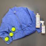 Eine blaue Steppjacke liegt flach auf einer grauen Oberfläche, begleitet von drei gelben Tennisbällen und zwei Reinigungsmittelflaschen. Das Arrangement hebt die leuchtenden Farben vor dem neutralen Hintergrund hervor.