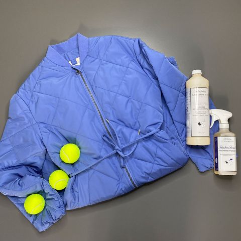 Eine blaue Steppjacke liegt flach auf einer grauen Oberfläche, begleitet von drei gelben Tennisbällen und zwei Reinigungsmittelflaschen. Das Arrangement hebt die leuchtenden Farben vor dem neutralen Hintergrund hervor.