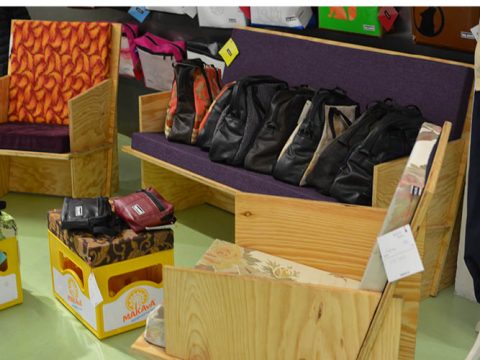 Eine Ausstellung mit Holzmöbeln, darunter eine Bank mit violettem Kissen und ein Sessel mit orangefarbenem Muster. Auf den Möbeln stehen mehrere Ledertaschen, weitere Taschen liegen in einem Karton auf dem Boden. Im Hintergrund hängen verschiedene Gegenstände.