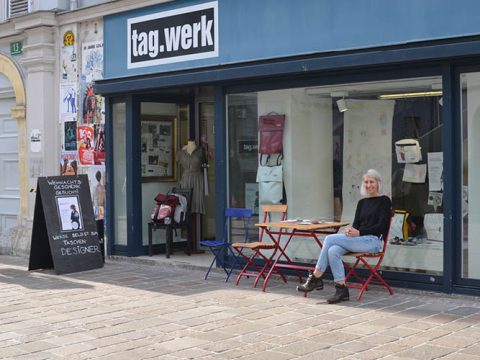 Eine Person sitzt auf einem bunten Stuhl vor einem Laden namens „tag.werk“. Im Schaufenster des Ladens finden sich unter anderem Rucksäcke. Der Gehweg ist mit Kopfsteinpflaster ausgelegt, die Außenwand ist mit Plakaten geschmückt.