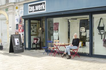 Eine Person sitzt an einem roten Tisch vor dem tag.werk-Laden. Im Schaufenster sind verschiedene Taschen ausgestellt. Die Fassade des Ladens hat weiße und blaue Wände. Im Inneren ist eine Schaufensterpuppe zu sehen. An der Wand links hängen Plakate.