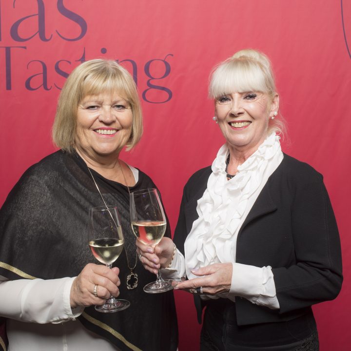 Zwei Frauen stehen lächelnd und mit Gläsern Weißwein in der Hand vor einem roten Hintergrund mit der Aufschrift „Glas Tasting“. Beide tragen elegante schwarz-weiße Outfits.
