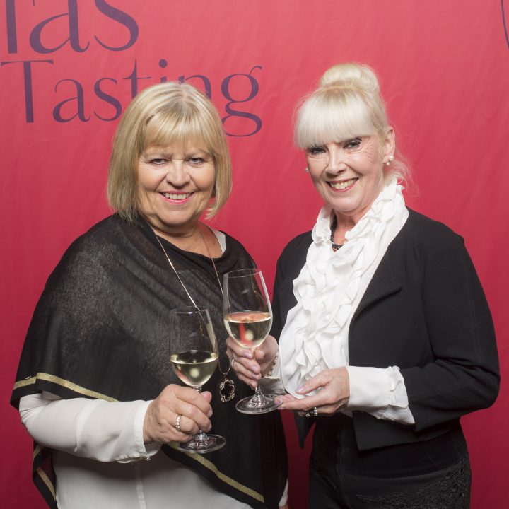 Zwei Frauen stehen lächelnd vor einem roten Hintergrund mit der Aufschrift „Glass Tasting“. Sie halten Gläser mit Weißwein in den Händen und sind in elegante schwarz-weiße Outfits gekleidet.