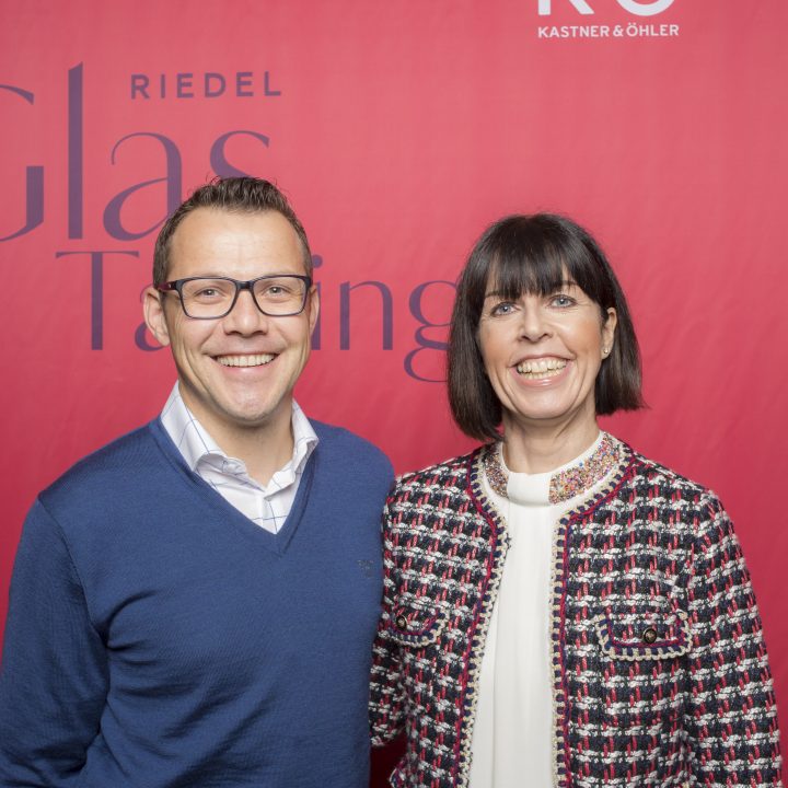 Ein Mann und eine Frau stehen vor einem roten Hintergrund mit der Aufschrift „Riedel Glas Tasting“. Der Mann trägt eine Brille, einen blauen Pullover und ein weißes Hemd, während die Frau eine gemusterte Jacke und eine weiße Bluse trägt. Beide lächeln.