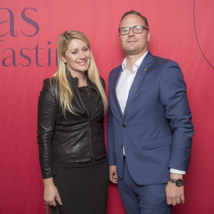 Ein Mann im blauen Anzug und eine Frau im schwarzen Outfit stehen lächelnd vor einem roten Hintergrund, auf dem die Worte „Glas Tasti“ teilweise sichtbar sind.