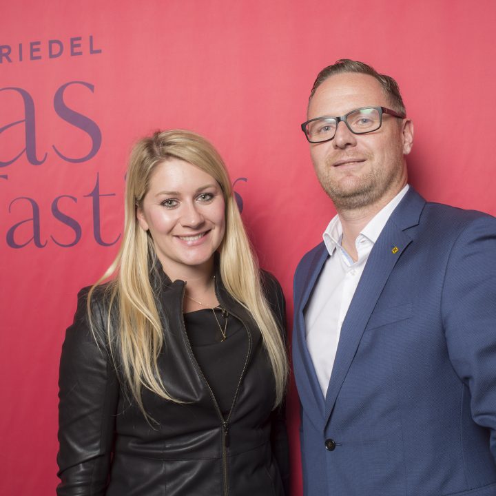Eine Frau mit langen blonden Haaren und einer schwarzen Lederjacke und ein Mann im blauen Anzug mit Brille stehen lächelnd vor einem roten Hintergrund mit der Aufschrift „Riedel Glass Tasting“.