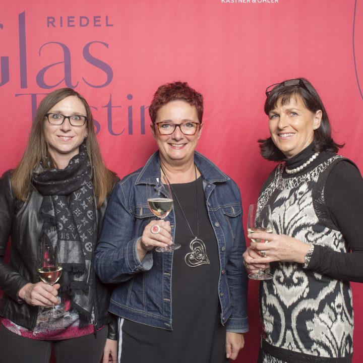 Drei Frauen stehen vor einem roten Hintergrund mit der Aufschrift „Riedel Glas Tasting“. Sie lächeln und halten Weingläser in den Händen. Die Frau in der Mitte hat rote Haare und trägt eine Brille, flankiert von Frauen mit braunem Haar.