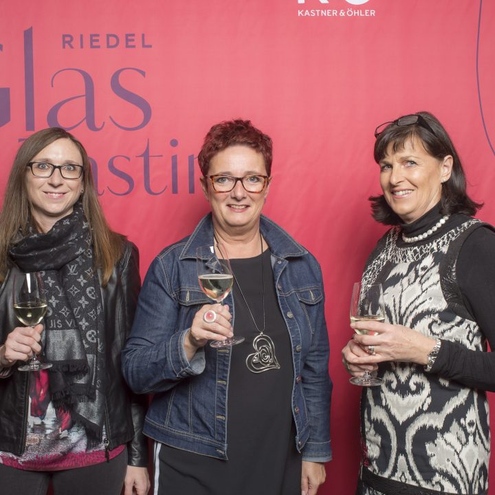 Drei Frauen stehen vor einem roten Hintergrund mit der Aufschrift „Glas“. Sie halten Gläser mit Weißwein in den Händen und lächeln in die Kamera. Sie sind lässig und elegant gekleidet.