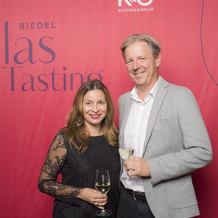 Eine Frau im schwarzen Spitzentop und ein Mann im hellgrauen Blazer lächeln und halten Weingläser in den Händen. Sie stehen vor einem roten Hintergrund mit dem Text „Riedel Glas Tasting“.