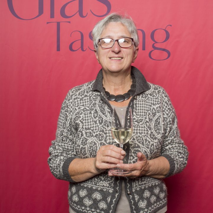 Eine ältere Person mit Brille und gemusterter Jacke hält ein Glas Wein und steht vor einem roten Hintergrund mit der Aufschrift „Riedel Glas Tasting“.