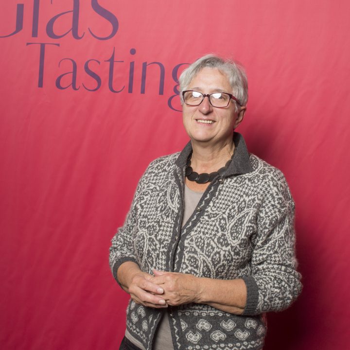 Eine Person mit kurzen grauen Haaren und Brille steht vor einem roten Hintergrund mit der Aufschrift „Riedel Glas Tasting“. Sie trägt einen gemusterten Pullover und einen schwarzen Rock.