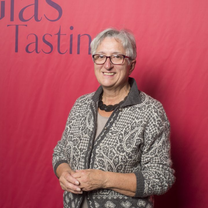 Eine Person mit kurzen grauen Haaren, Brille, gemustertem Pullover und schwarzem Stepprock steht vor einem roten Hintergrund mit den Aufschriften „Riedel Glas Tasting“ und „Kastner & Öhler“. Sie lächeln und haben die Hände gefaltet.