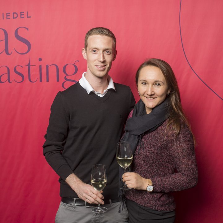Ein Mann und eine Frau stehen lächelnd und mit Weißweingläsern in der Hand vor einem roten Hintergrund mit der Aufschrift „Riedel Glas Tasting“.