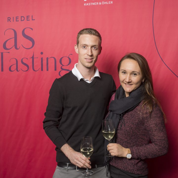 Ein Mann und eine Frau stehen vor einem roten Hintergrund mit der Aufschrift „Riedel Glas Tasting“. Sie lächeln und halten Gläser mit Weißwein in den Händen. Die Frau trägt einen Schal und der Mann einen Pullover.