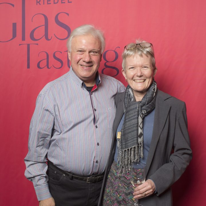 Ein lächelnder Mann und eine lächelnde Frau posieren vor einem roten Hintergrund und werben für eine Glasverkostung von Riedel und Kastner & Öhler. Der Mann trägt ein gestreiftes Hemd, die Frau hat einen Schal und hält ein Weinglas.