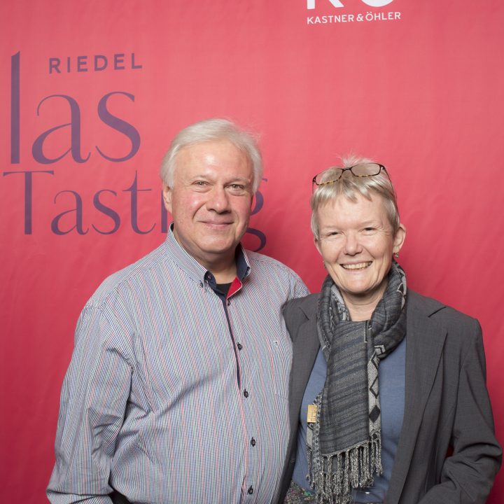 Ein lächelndes Paar steht vor einem roten Hintergrund mit der Aufschrift „Riedel Glas Tasting“. Der Mann trägt ein gestreiftes Hemd, die Frau ein Kopftuch und eine Brille auf dem Kopf.