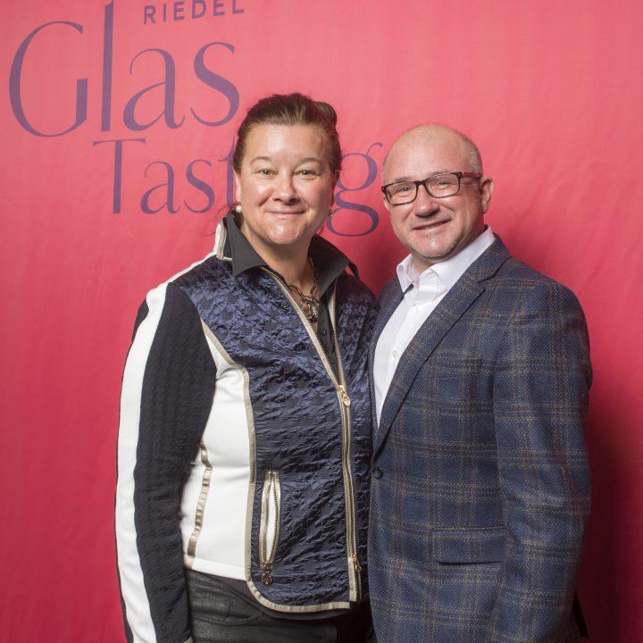 Eine Frau und ein Mann stehen lächelnd vor einem roten Hintergrund mit den Aufschriften „Riedel Glas Tasting“ und „Kastner & Öhler“. Die Frau trägt eine Weste, der Mann einen karierten Blazer.