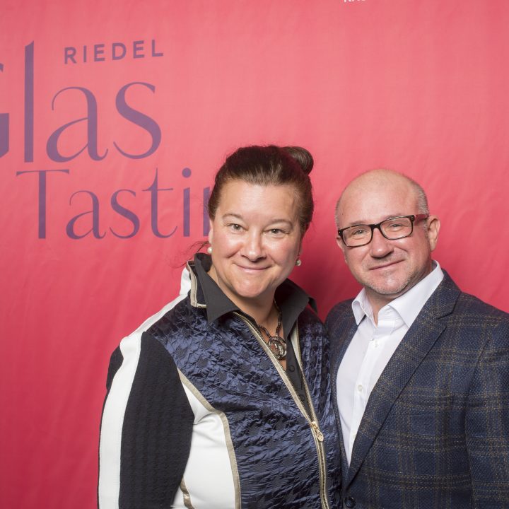 Eine Frau und ein Mann stehen lächelnd vor einem roten Hintergrund mit der Aufschrift „Riedel Glas Tasting“. Die Frau trägt eine schwarz-weiße Jacke, der Mann eine karierte Jacke und ein weißes Hemd.