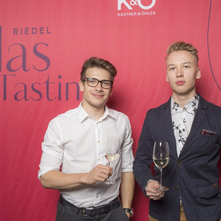 Zwei Männer stehen vor einem roten Hintergrund mit der Aufschrift „Riedel Glas Tasting“. Beide halten Weingläser in den Händen. Der Mann links trägt ein weißes Hemd und eine Brille, der rechte einen dunklen Blazer über einem gemusterten Hemd.