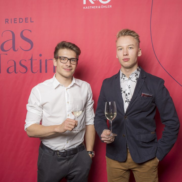 Zwei Personen stehen vor einem roten Hintergrund mit der Aufschrift „Riedel Glas Tasting“. Sie halten Weingläser in der Hand und tragen elegante Freizeitkleidung; einer trägt ein weißes Hemd und der andere einen dunklen Blazer.