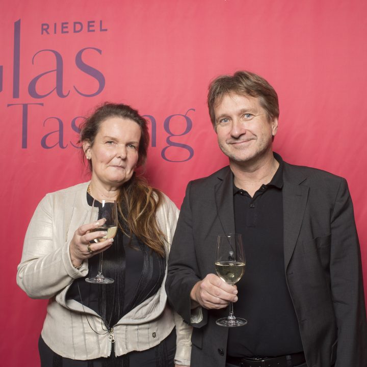 Ein Mann und eine Frau in Abendgarderobe stehen vor einem roten Hintergrund mit der Aufschrift „Riedel Glas Tasting“ und halten jeweils ein Glas Weißwein in der Hand.