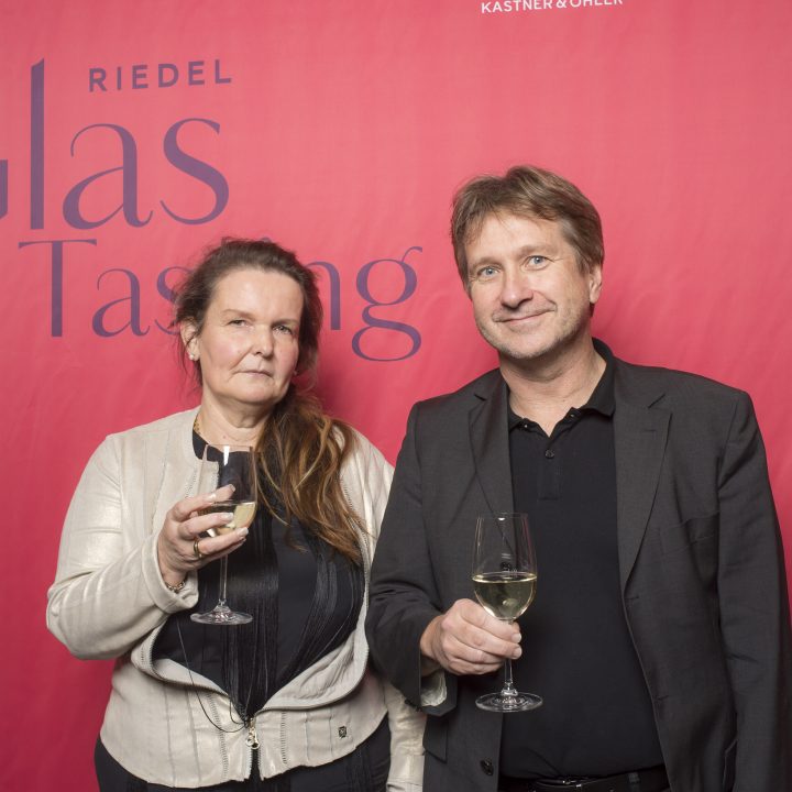 Vor einem roten Hintergrund mit der Aufschrift „Riedel Glass Tasting“ stehen zwei Personen und halten Weingläser in den Händen. Die Frau trägt eine helle Jacke, der Mann einen dunklen Anzug. Beide blicken in die Kamera.