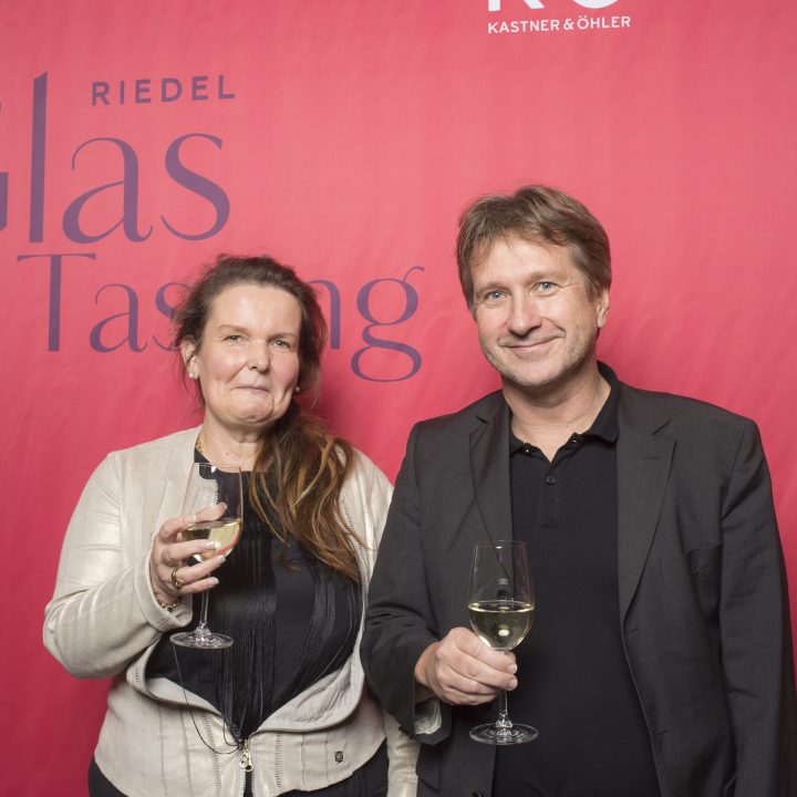Zwei Personen stehen mit Weingläsern vor einem roten Hintergrund mit dem Text „Riedel Glas Tasting“. Beide lächeln, wobei die Frau links eine helle Jacke und der Mann rechts einen dunklen Anzug trägt.