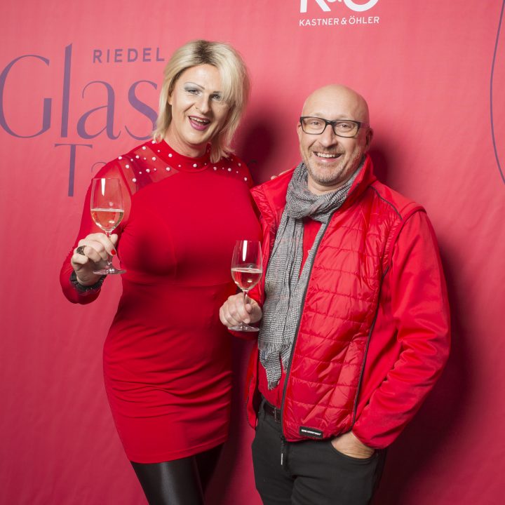 Zwei Personen lächeln und halten Weingläser vor einem rosa Hintergrund mit den Logos „Riedel Glas“ und „Kastner & Öhler“. Eine Person trägt ein rotes Kleid, die andere eine rote Jacke und einen grauen Schal.