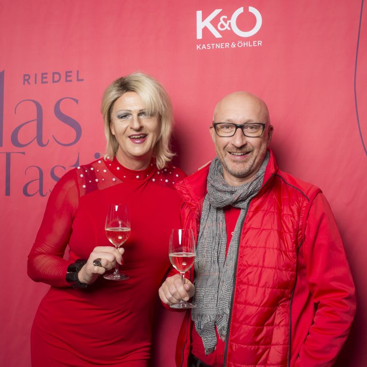 Zwei Personen stehen lächelnd vor einem roten Hintergrund mit den Logos „Riedel Glas Tasting“ und „Kastner & Öhler“. Beide halten Gläser mit Weißwein in den Händen und tragen rote Kleidung.