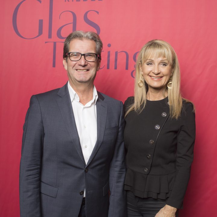 Ein Mann und eine Frau stehen vor einem roten Hintergrund, auf dem die Worte „Riedel Glas Tasting“ und „K&Ö Kastner & Öhler“ zu sehen sind. Der Mann trägt einen dunklen Anzug und die Frau ein schwarzes Oberteil. Beide lächeln in die Kamera.