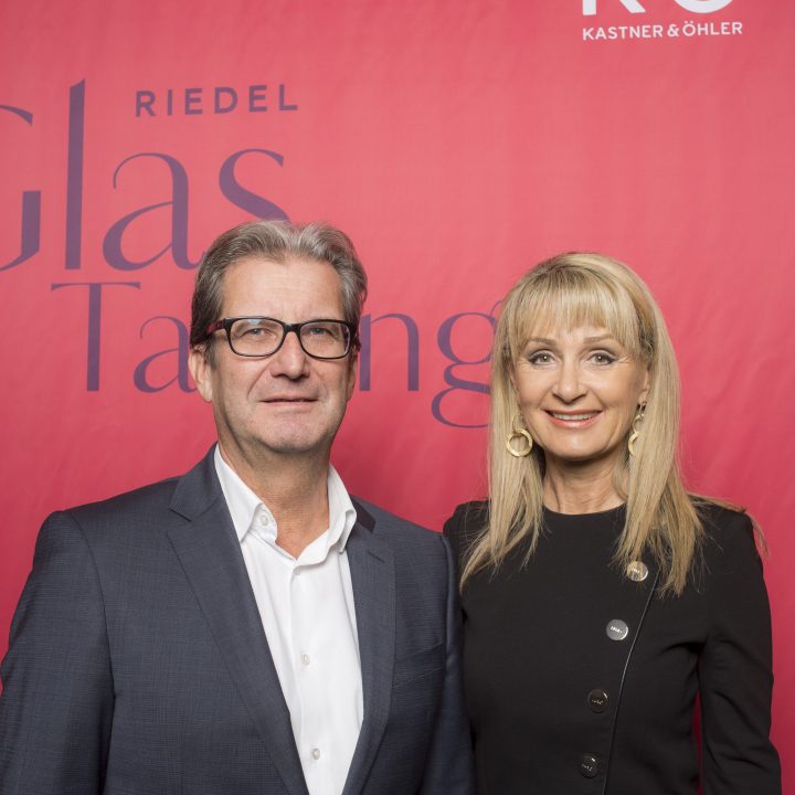 Ein Mann in dunklem Anzug und Brille steht neben einer Frau mit langen blonden Haaren und schwarzem Outfit. Sie stehen vor einem roten Hintergrund mit teilweise sichtbarem Text.