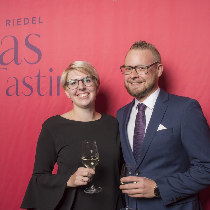 Eine Frau im schwarzen Kleid und ein Mann im blauen Anzug stehen lächelnd und halten Weingläser vor einem roten Hintergrund mit dem Text „Riedel Glas Tasting“.