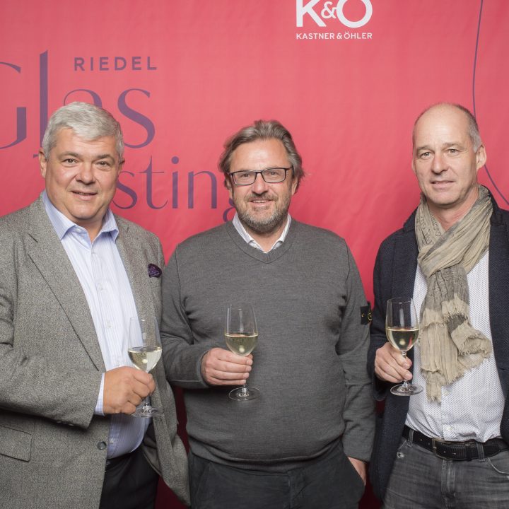 Drei Männer stehen nebeneinander vor einem roten Hintergrund und halten Weingläser in den Händen. Sie tragen legere Geschäftskleidung und scheinen an einer Weinveranstaltung teilzunehmen.