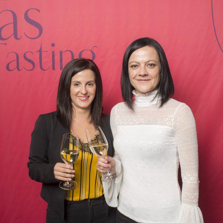 Vor einem roten Hintergrund mit der Aufschrift „Glas Tasting“ stehen zwei lächelnde Menschen mit Weingläsern in den Händen. Eine trägt ein gelbes Oberteil unter einer schwarzen Jacke, die andere eine weiße Bluse.